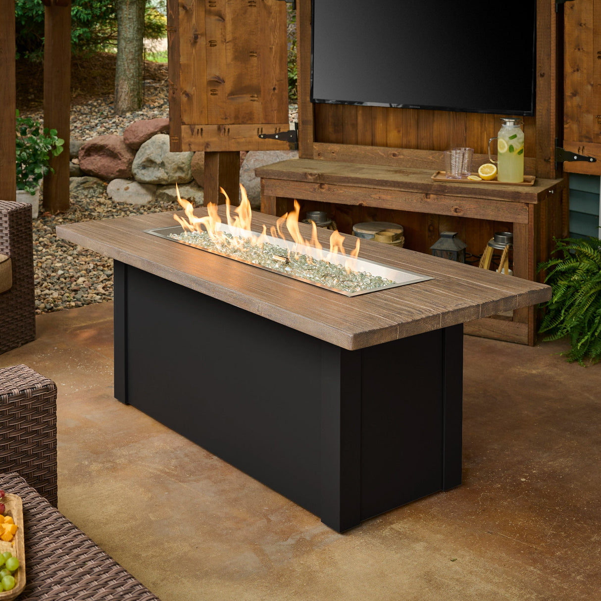The Havenwood Linear Gas Fire Pit Table with a Driftwood top and Luverne Black base in a patio setting next to a large TV