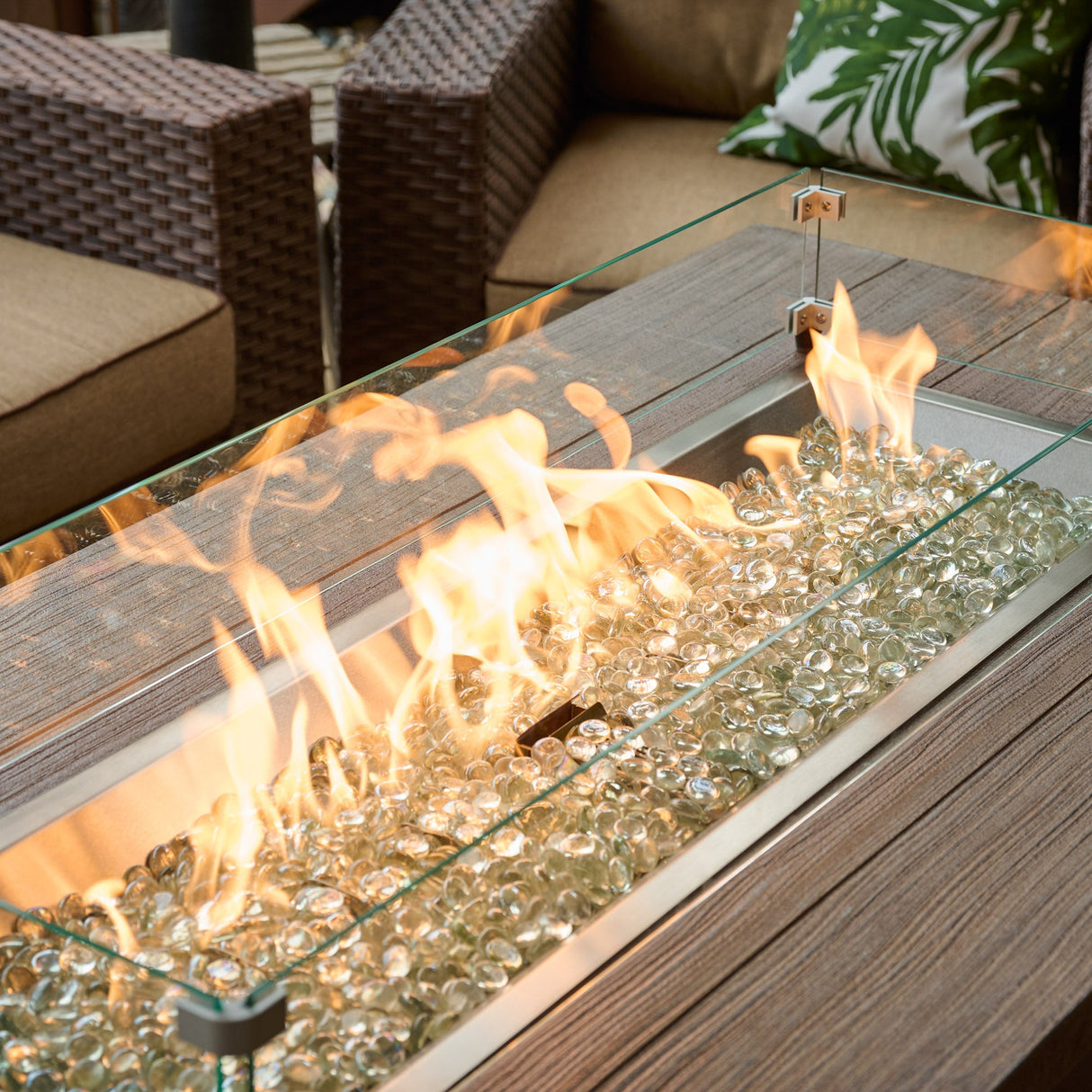 A glass wind guard protecting the flame coming from a Havenwood Linear Gas Fire Pit Table with a Driftwood top and Luverne Black base