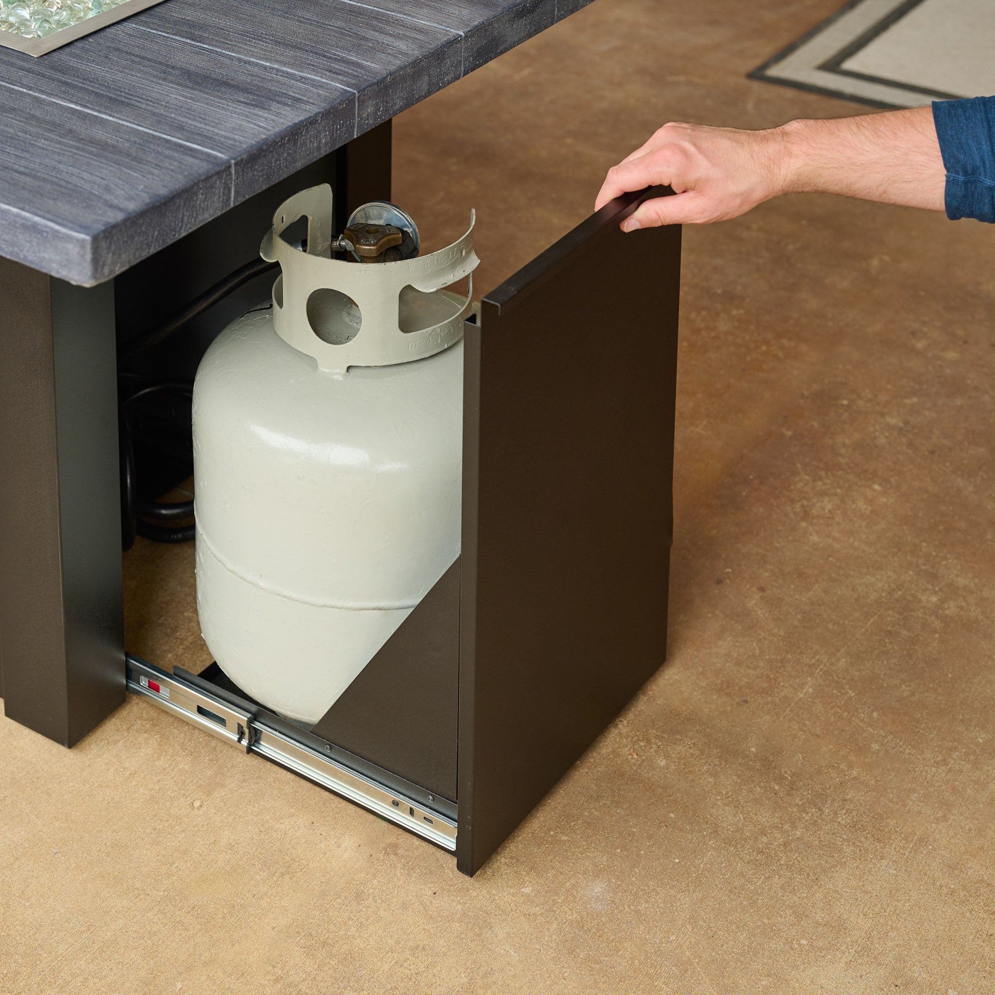 An individual opening the side access door on a Havenwood Linear Gas Fire Pit Table with a Carbon Grey top and Luverne Black base