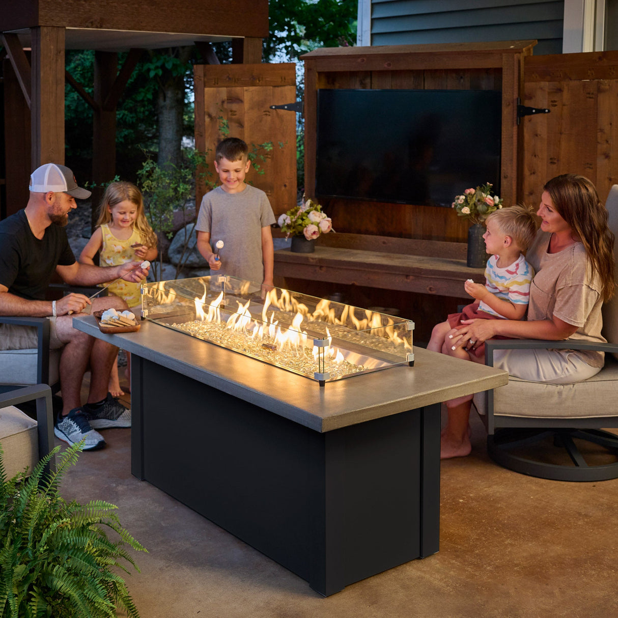 A family gathered around the Havenwood Linear Gas Fire Pit Table with a Pebble Grey top and Luverne Black base roasting marshmallows