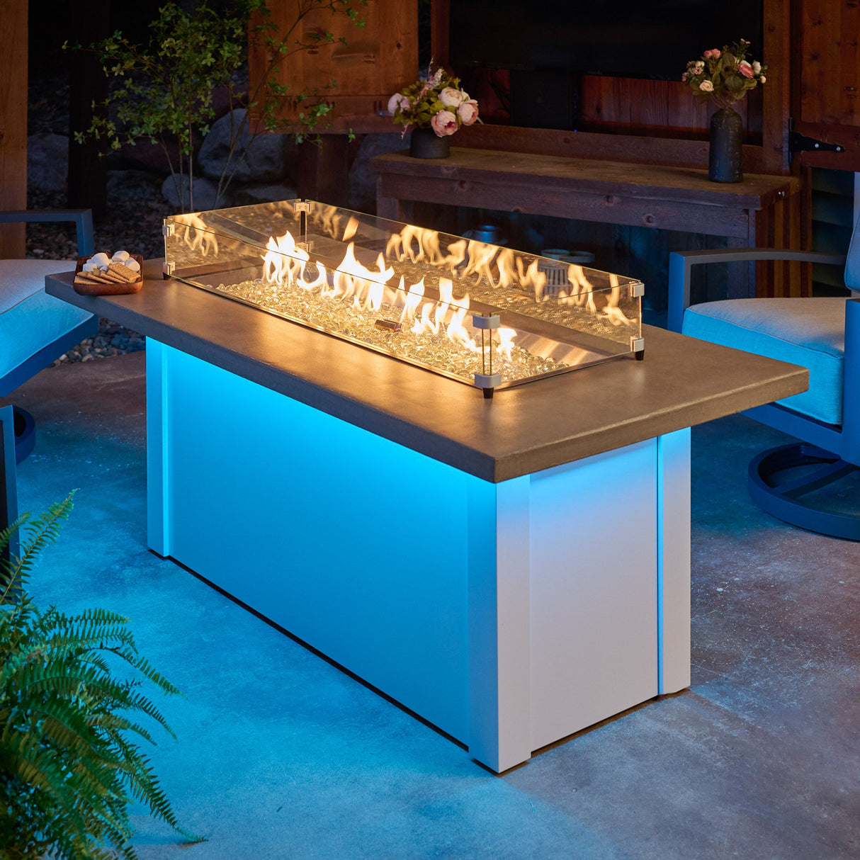 The Havenwood Linear Gas Fire Pit Table with a Pebble Grey top and White base being used on a dark night in a patio setting