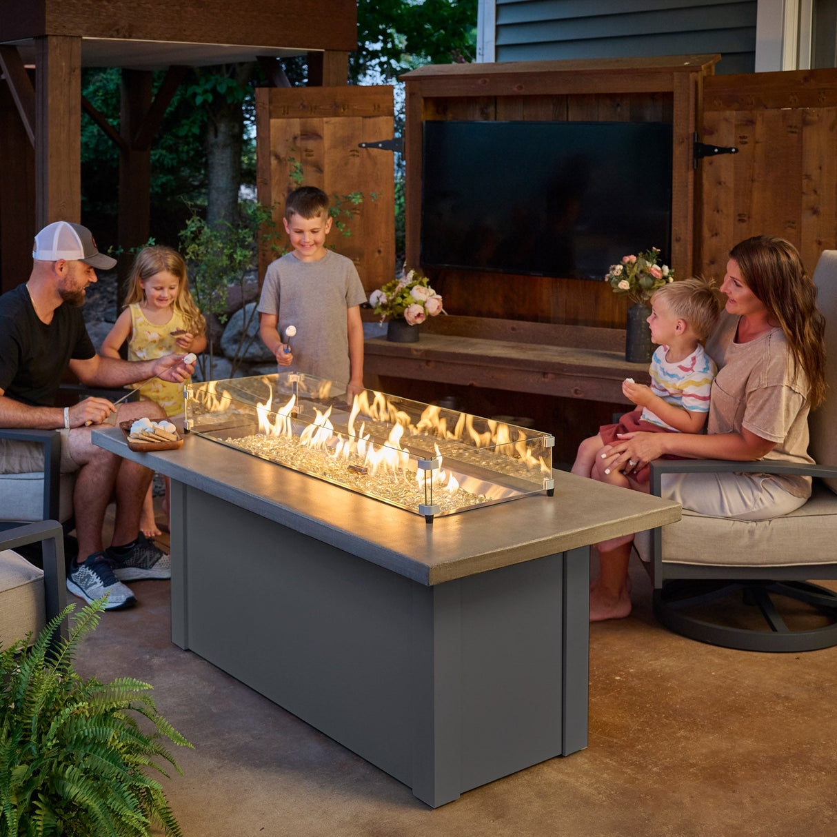 A family gathered around the Havenwood Linear Gas Fire Pit Table with a Pebble Grey top and Graphite Grey base roasting marshmallows