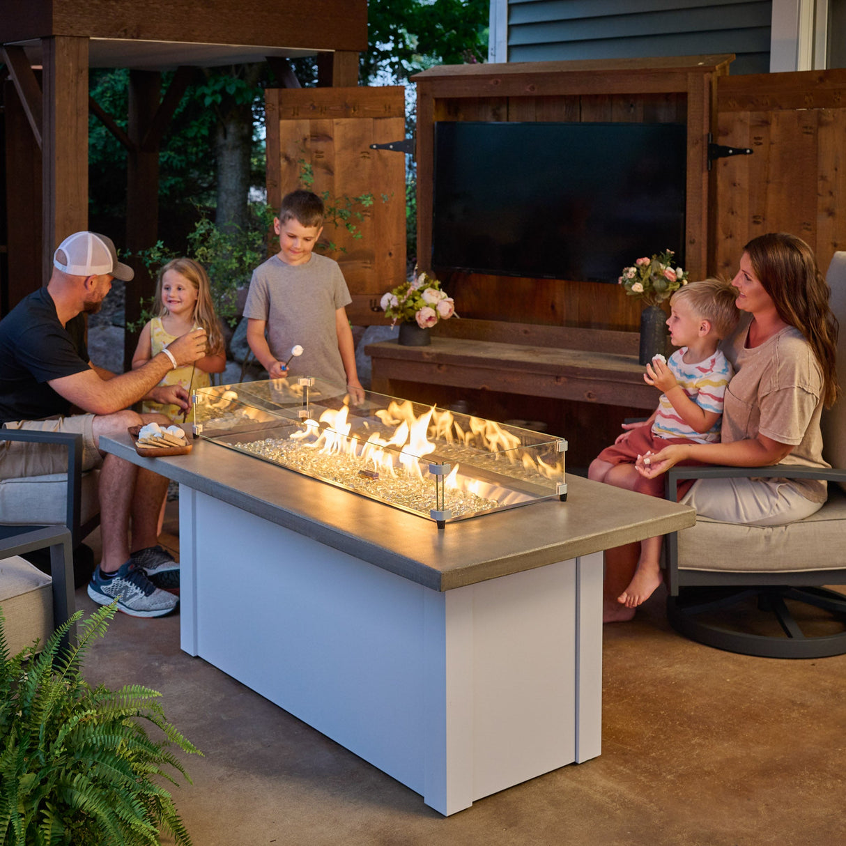 A family gathered around the Havenwood Linear Gas Fire Pit Table with a Pebble Grey top and White base roasting marshmallows