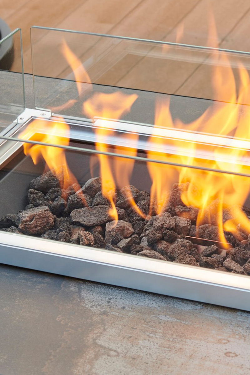 A close up view of a folding wind guard and lava rock being used on a linear gas fire pit table