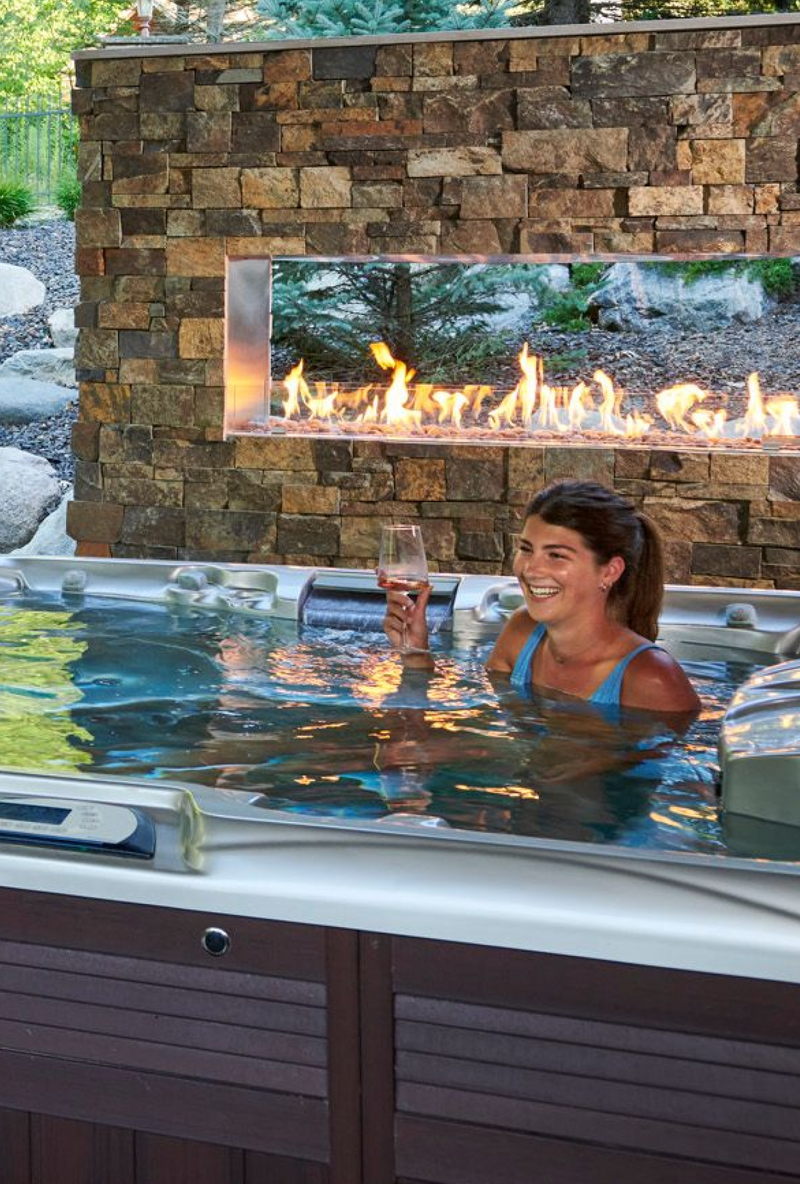 An individual relaxing in a hot tub next to a See Through Ready to Finish Outdoor Fireplace on a patio