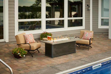An image of a Cedar Ridge Gas Fire Pit Table on a patio. It's burning and has a pair of drinks on one corner. The fire pit table is positioned in front of two patio chairs near a pool.