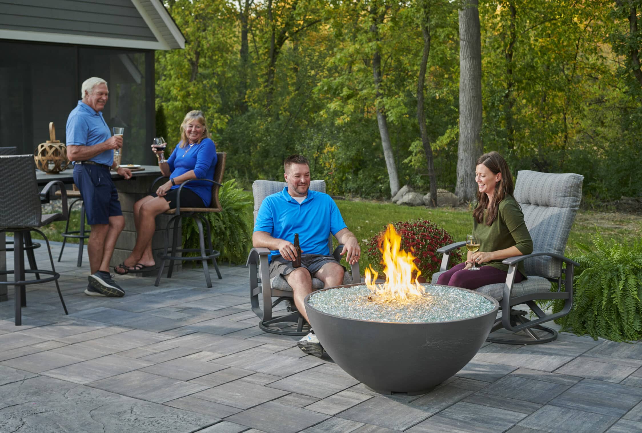 Friends gathered around a round gas fire pit on a backyard patio surrounded by trees.