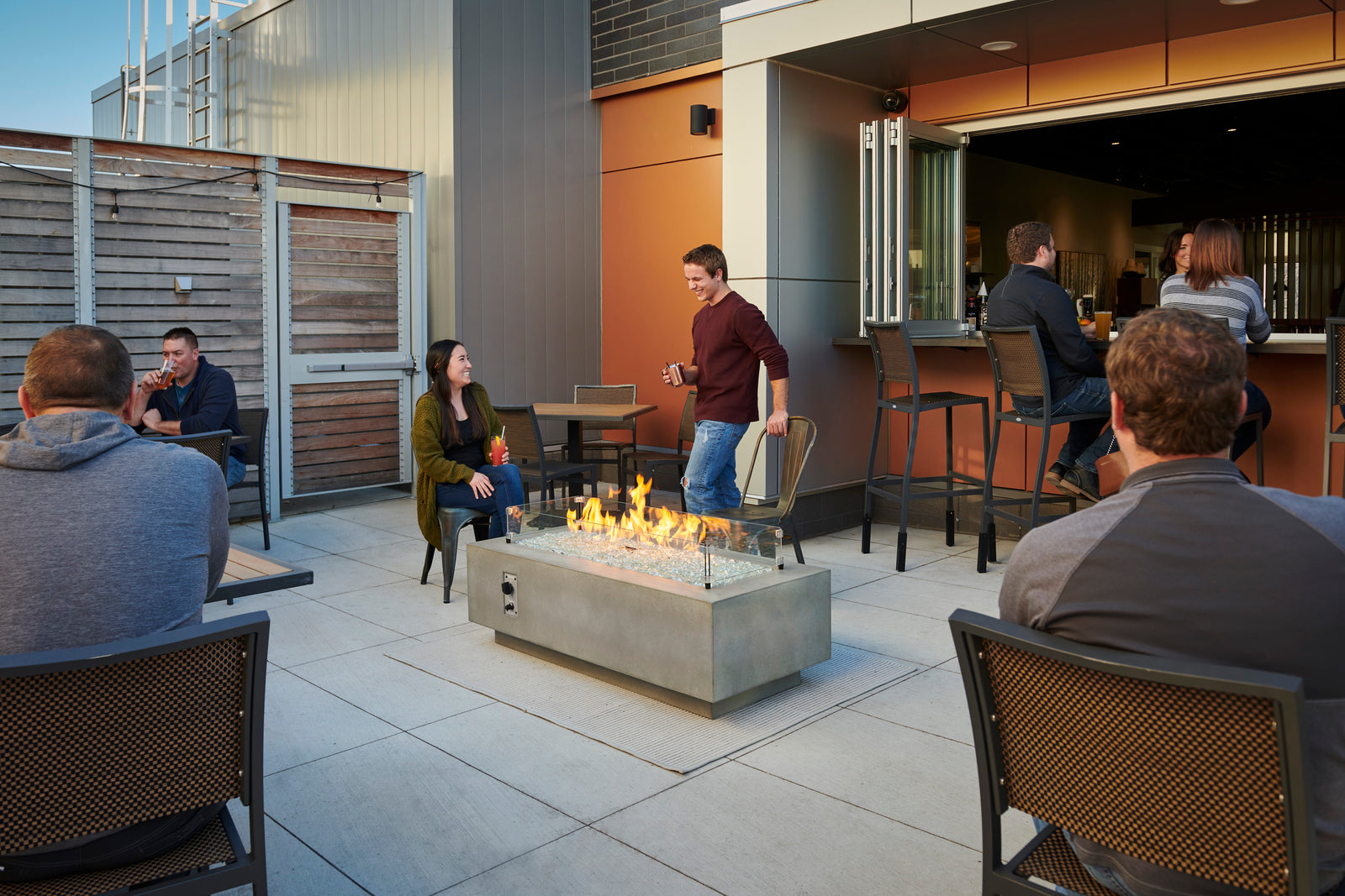 A man and a woman take a seat around a Cove 54" rectangular gas fire pit. They're on a patio a restaurant.