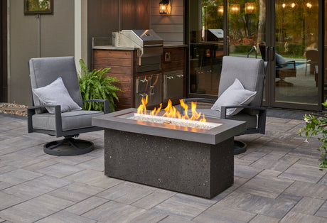 A Key Largo Gas Fire Pit by The Outdoor GreatRoom Company. It's on a patio surrounded by patio chairs. An outdoor kitchen and and overhead outdoor decorative lights can be seen in the image as well.