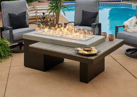 An image of the Uptown Coastal Grey gas fire pit table. It's flames are very visible and protected by a glass guard. There's a plate of chips and salsa on the front corner of the table. There are patio chairs, a pool, and plants behind the table.