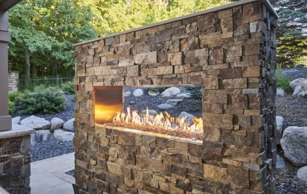 The See Through Ready to Finish Outdoor Fireplace in an outdoor setting of trees and rocks