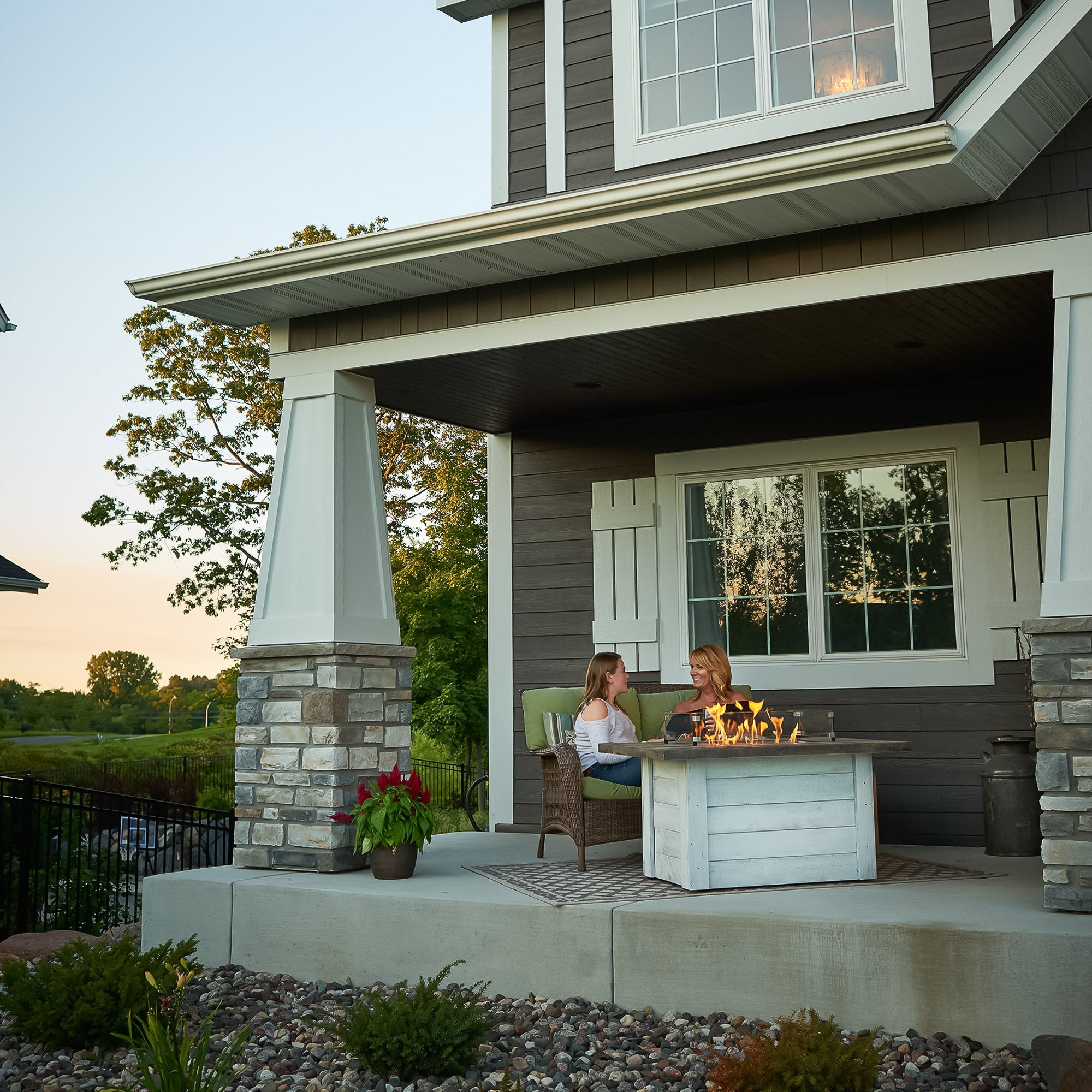 Two individuals relaxing next to the Alcott Rectangular Gas Fire Pit Table at the front of a house