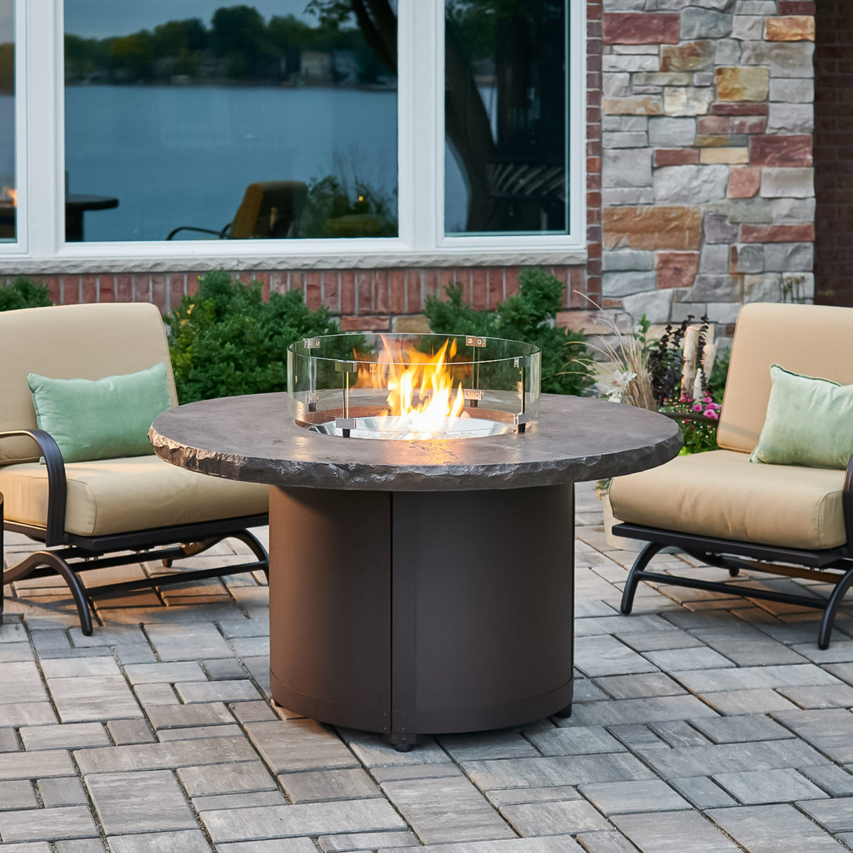 The Marbleized Noche Beacon Round Gas Fire Pit Table outside with a round glass wind guard protecting the flame