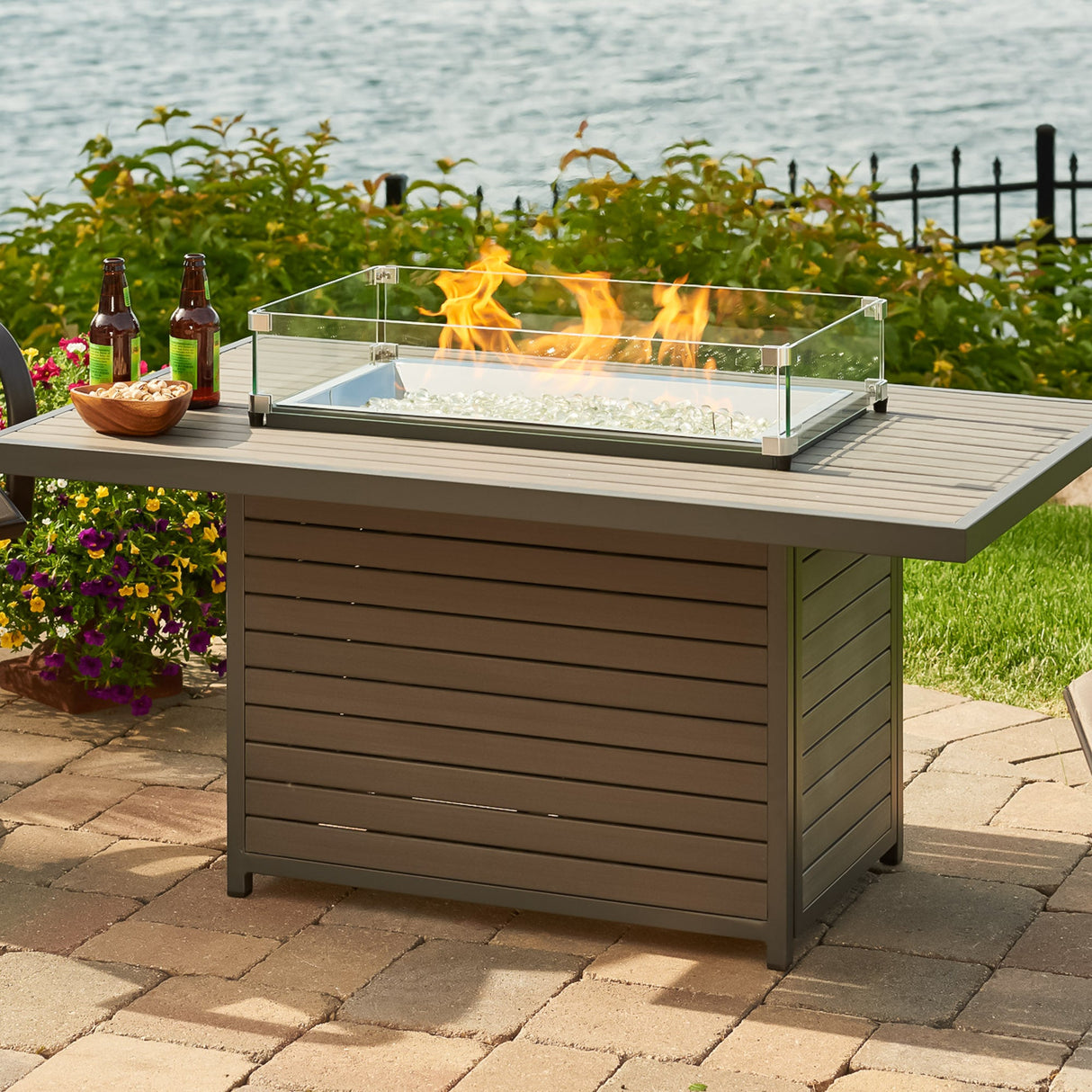 The Brooks Rectangular Gas Fire Pit Table next to a lake with food and drink placed on its top