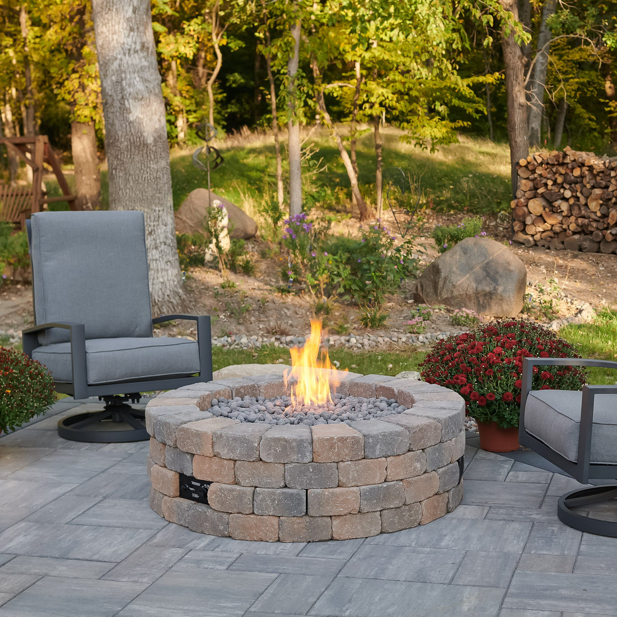 The Bronson Block Round Gas Fire Pit Kit surrounded by patio furniture and a forest background