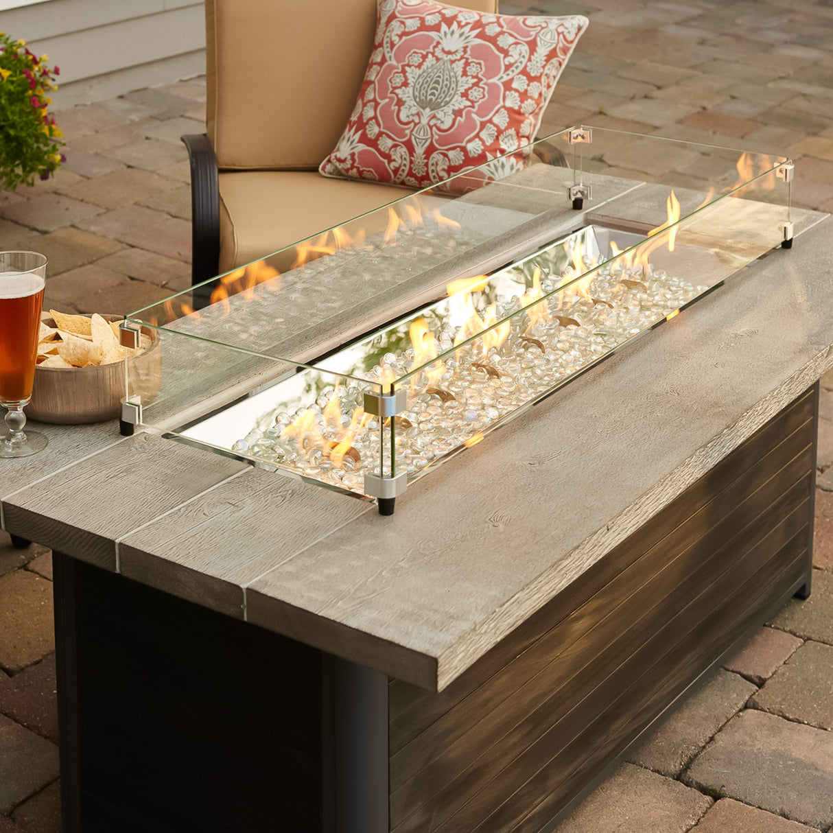 A close up view of the Cedar Ridge Linear Gas Fire Pit Table with a glass wind guard being used to protect the flame from the wind