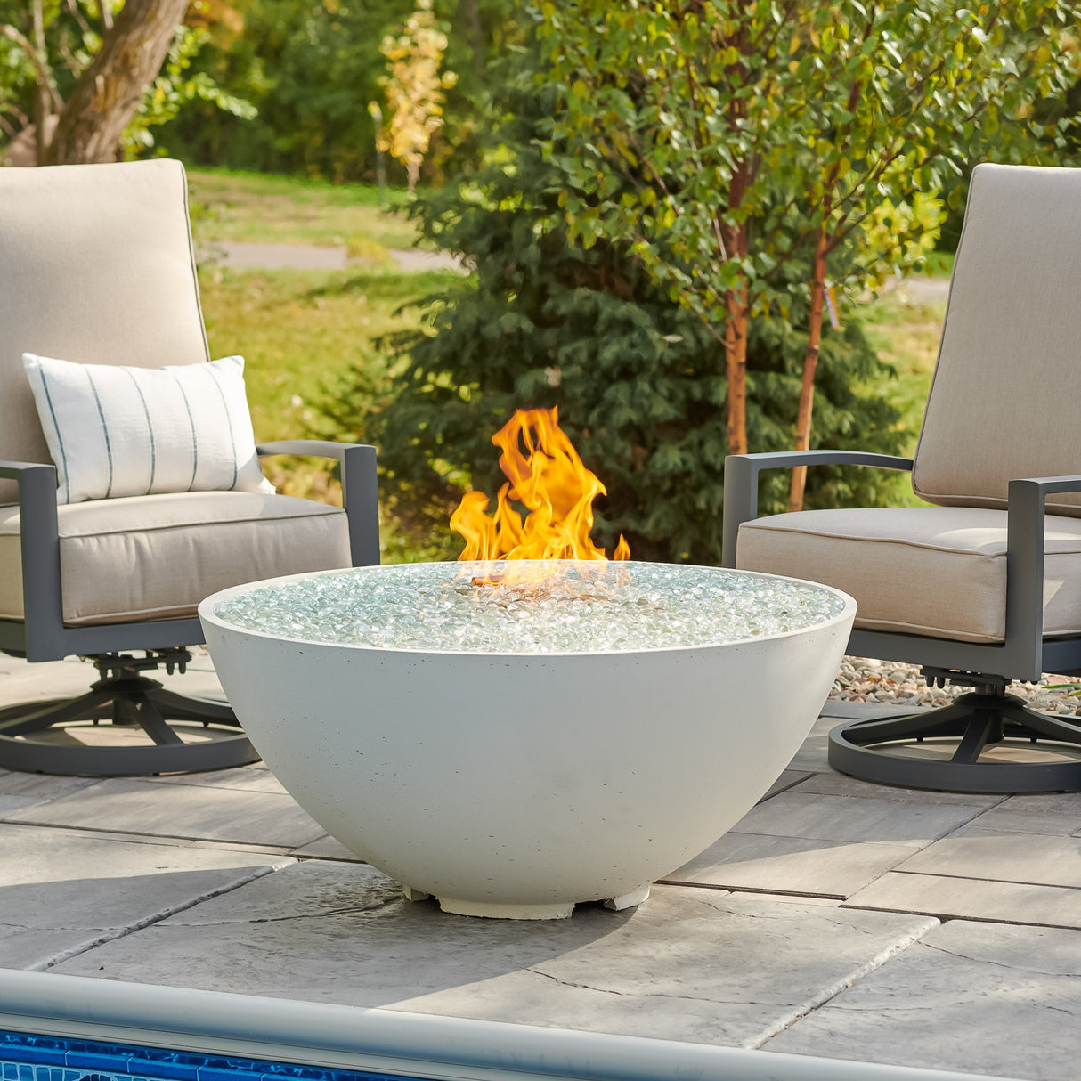 A large flame coming from the White Cove Edge Round Gas Fire Pit Bowl 42" surrounded by lounge chairs