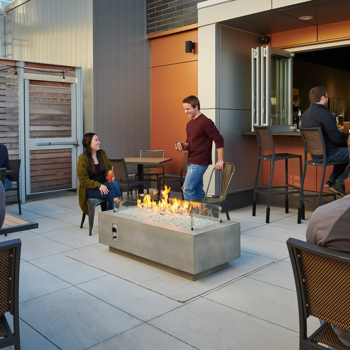A commercial restaurant space with a Natural Grey Cove Linear Gas Fire Pit Table 54" surrounded by two individuals