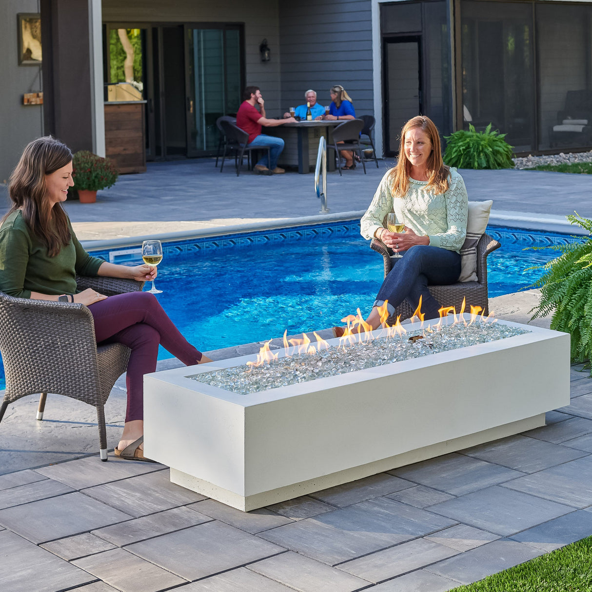 Individuals sitting around the White Cove Linear Gas Fire Pit Table 72" and relaxing next to the pool