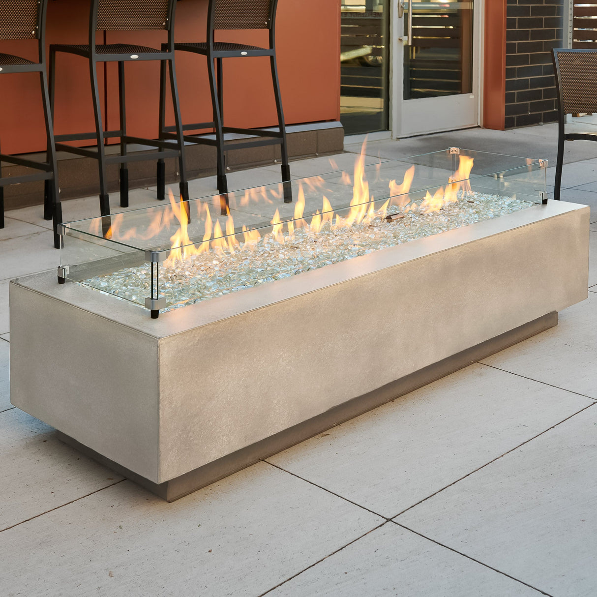 A glass wind guard placed on the top of a Natural Grey Cove Linear Gas Fire Pit Table 72" to protect the flame from the wind