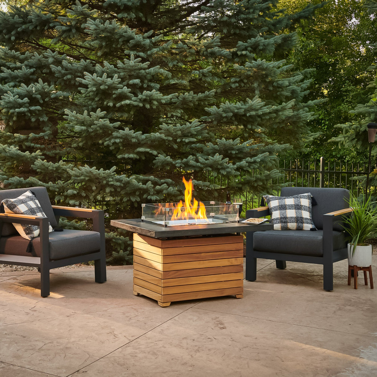 The Darien Rectangular Gas Fire Pit Table with an Everblend top and a glass wind guard next to patio furniture and a large pine tree