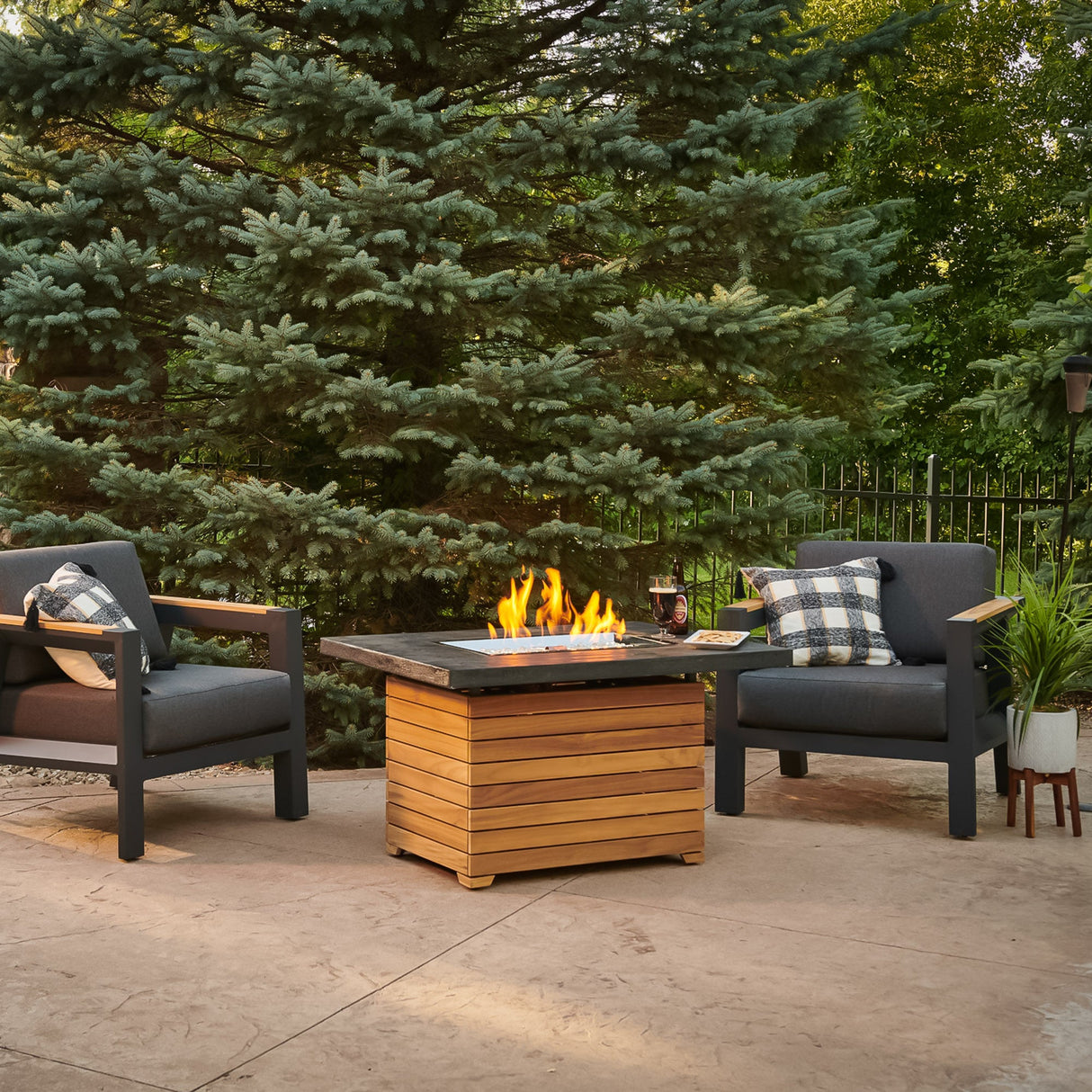 The Darien Rectangular Gas Fire Pit Table with an Everblend top outside next to pine trees and patio furniture