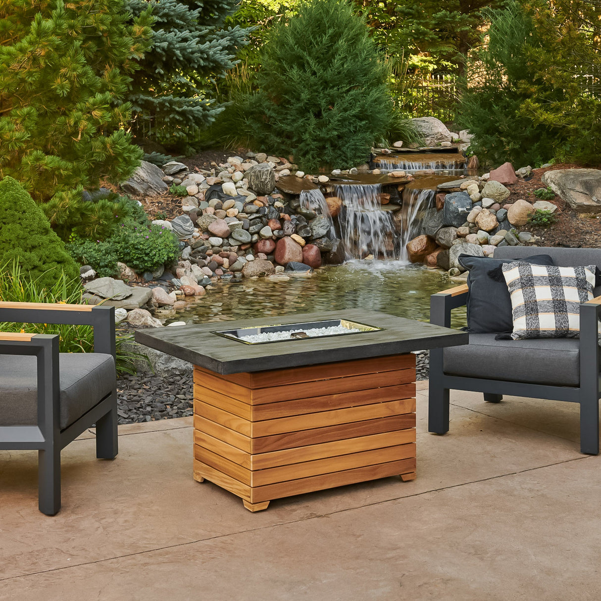 The Darien Rectangular Gas Fire Pit Table with an Everblend top next to a small waterfall and patio furniture