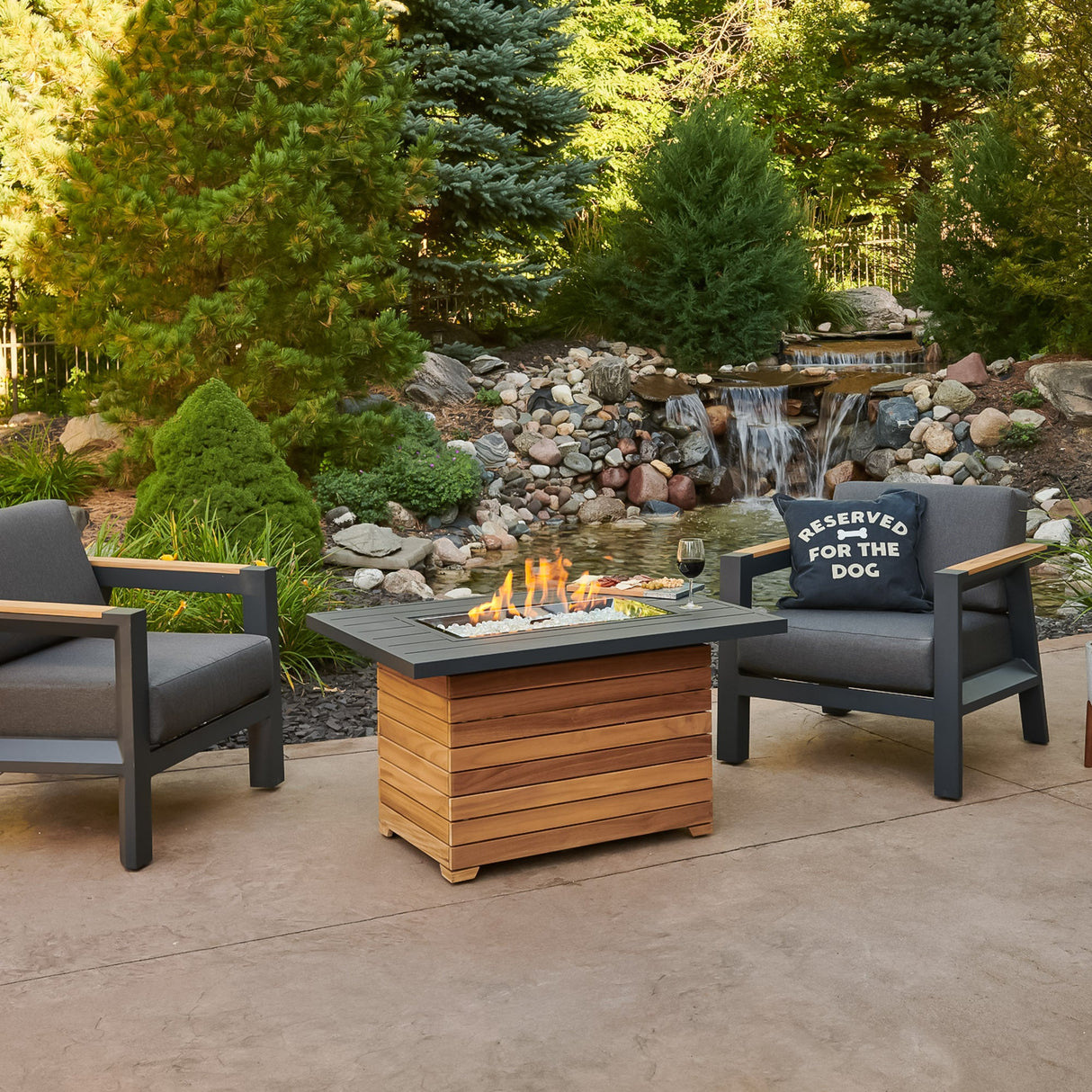 The Darien Rectangular Gas Fire Pit Table with an Aluminum top in a scenic outdoor setting with a waterfall and pine trees