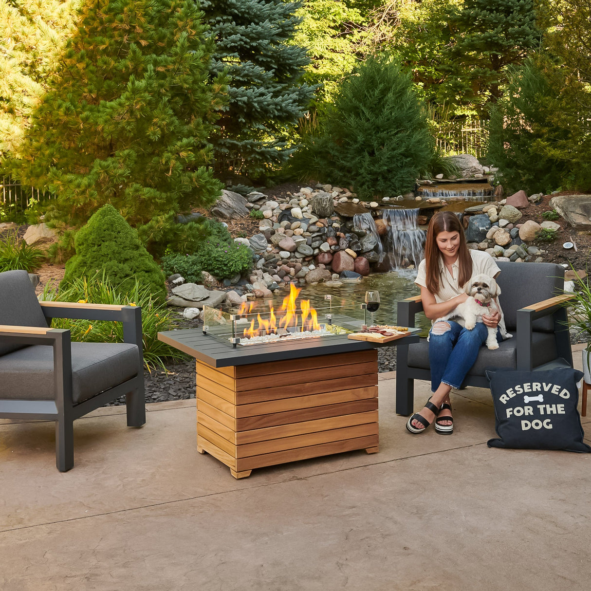 An individual and their dog relaxing next to the Darien Rectangular Gas Fire Pit Table with an Aluminum top