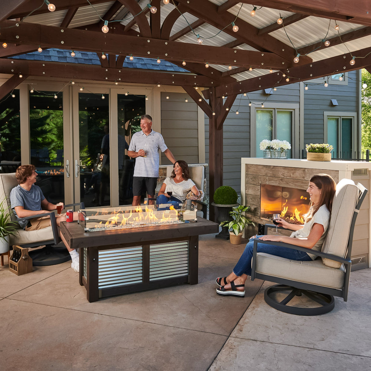 A group of individuals gathered around the Denali Brew Linear Gas Fire Pit Table under their pergola