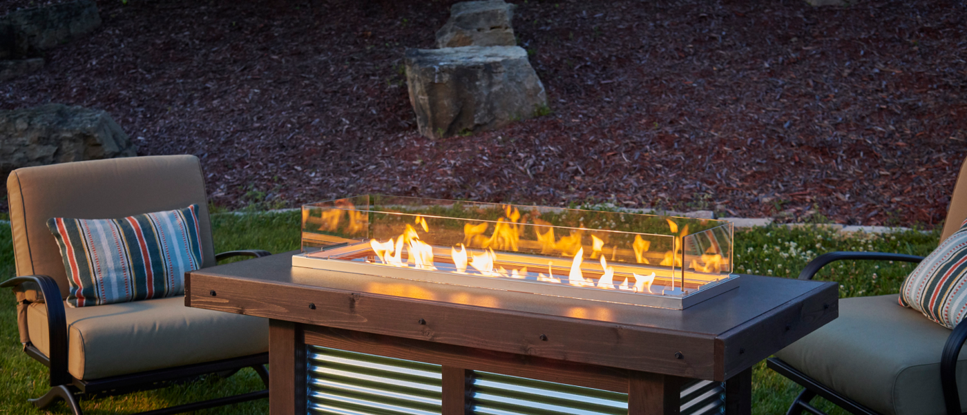 A Denali Brew Linear Gas Fire Pit Table being used on a backyard lawn with outdoor chairs surrounding the unit