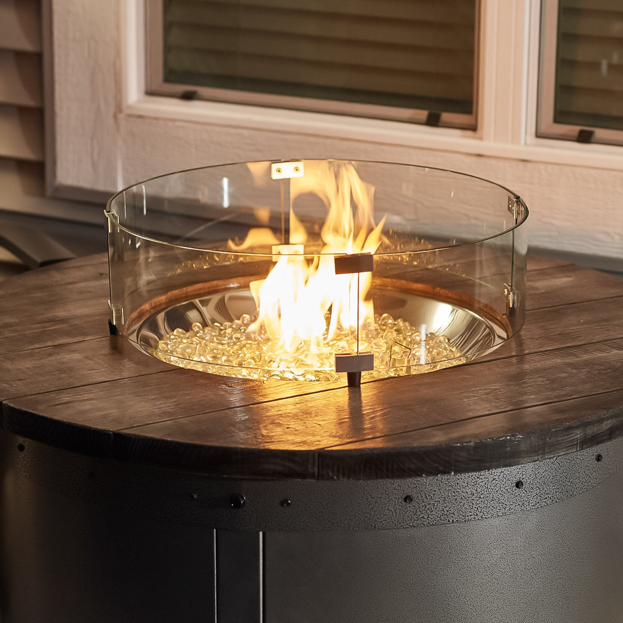 A glass wind guard placed on top of an Edison Round Gas Fire Pit Table, protecting the flame from the wind