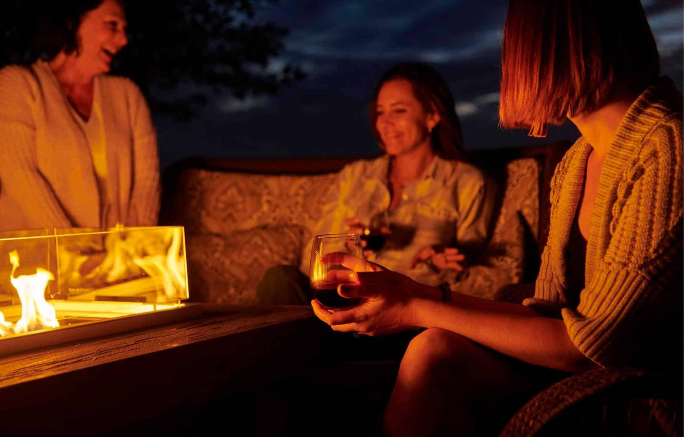 Three individuals having a conversation on a dark night around a linear gas fire pit table