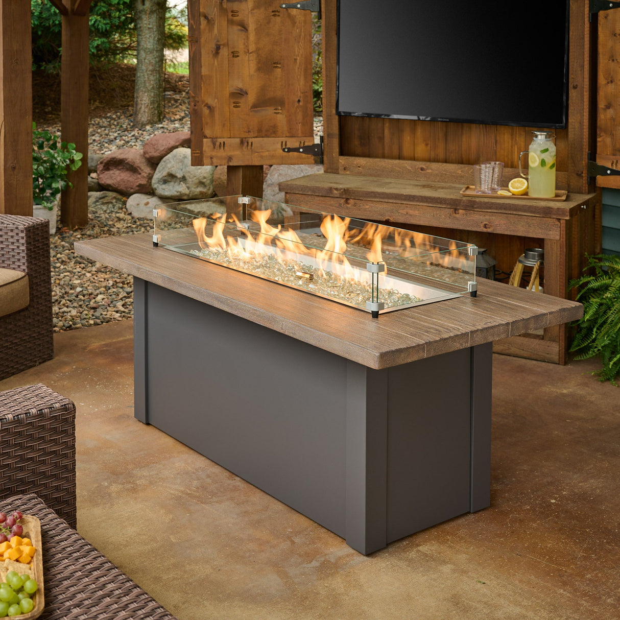 A glass wind guard protecting the flame coming from the burner of a Havenwood Linear Gas Fire Pit Table with a Driftwood top and Graphite Grey base