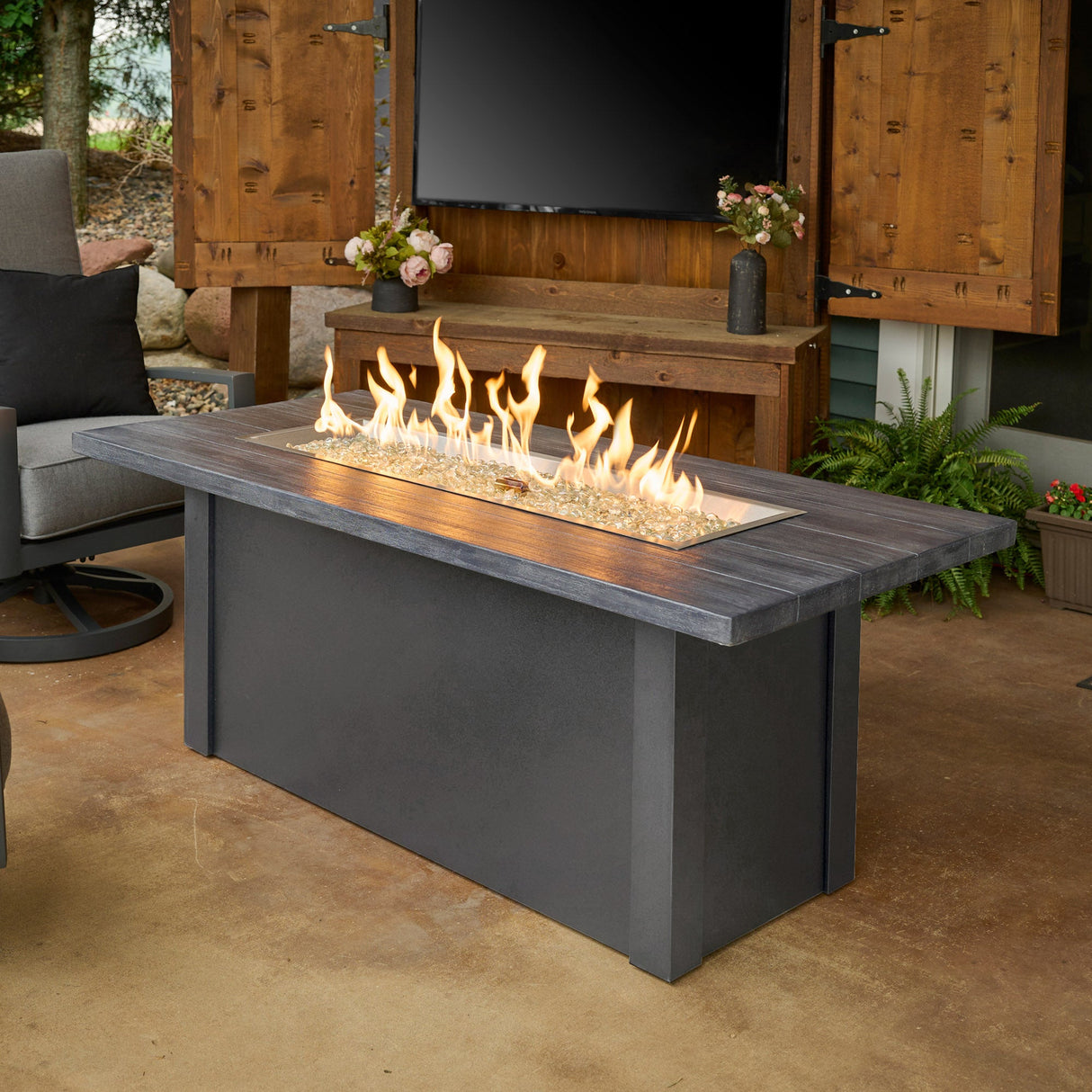 The Havenwood Linear Gas Fire Pit Table with a Carbon Grey top and Graphite Grey base in an outdoor space next to patio furniture