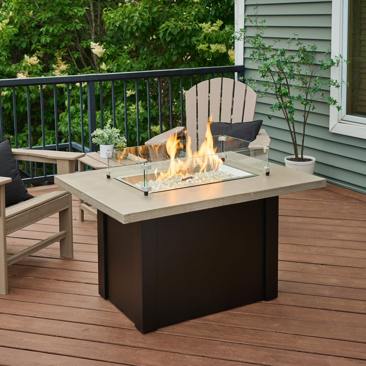 The Havenwood Rectangular Gas Fire Pit Table with a Pebble Grey top and Luverne Black base in an outdoor space with a glass wind guard on its top