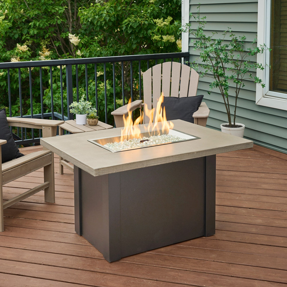 A patio setup that features a Havenwood Rectangular Gas Fire Pit Table with a Pebble Grey top and Graphite Grey base with a large flame