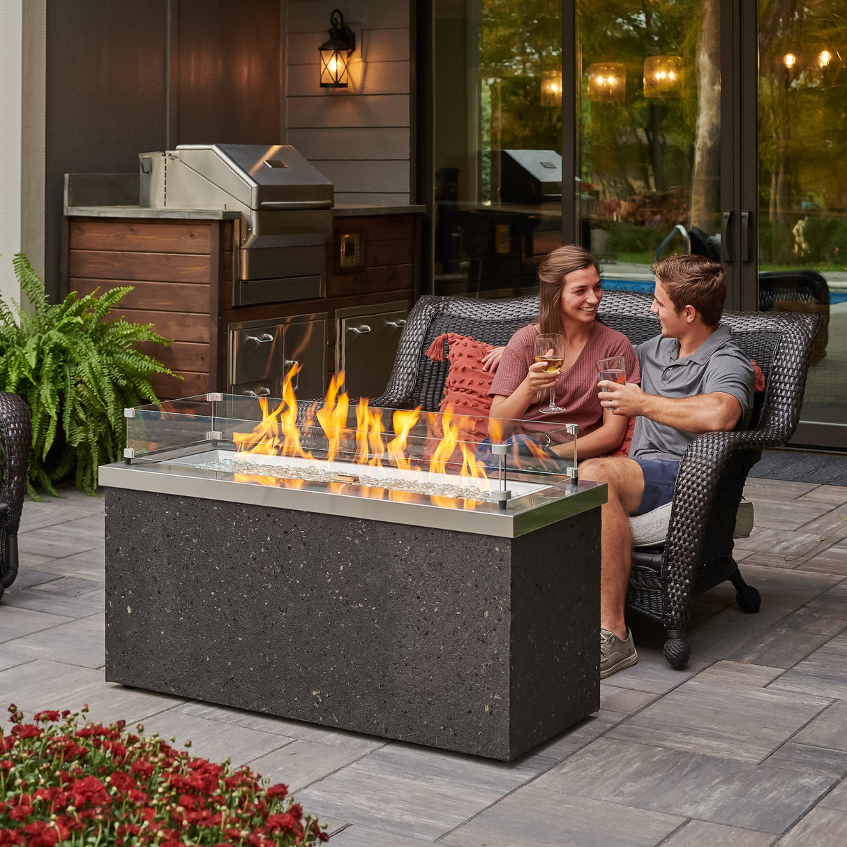 A couple sitting on a patio couch next to the Stainless Steel Key Largo Linear Gas Fire Pit Table
