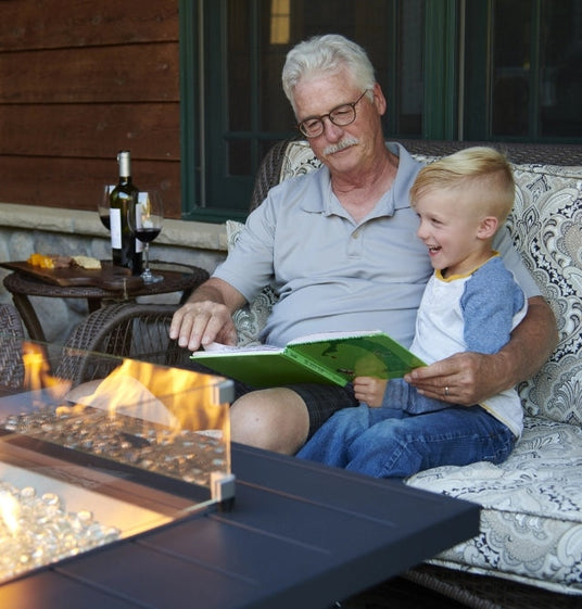 A young and old individual reading a book on an outdoor patio next to a fire pit table