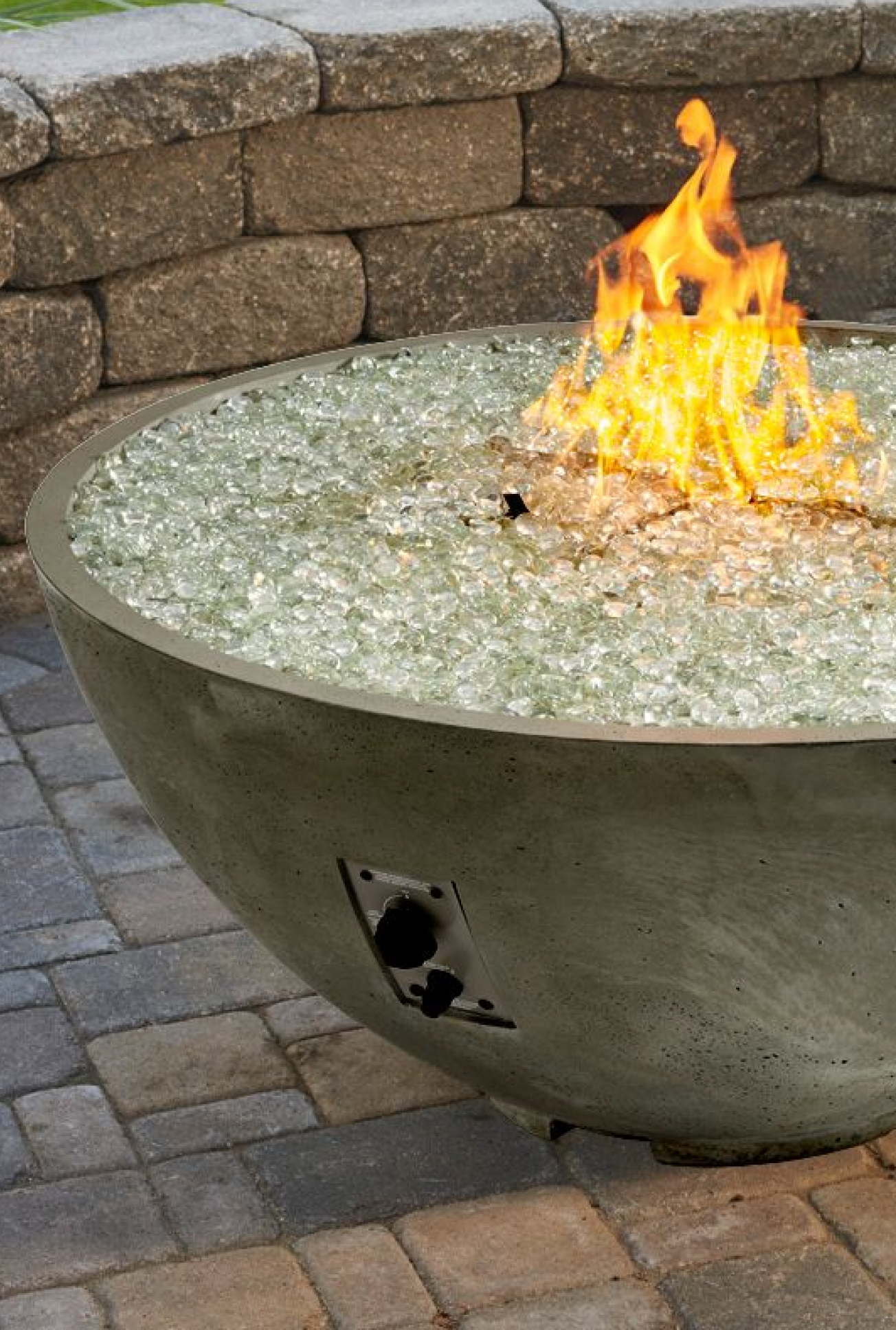 A close up view of temperred fire gems on the burner of a natural grey Cove Edge Round Gas Fire Pit Bowl on a patio