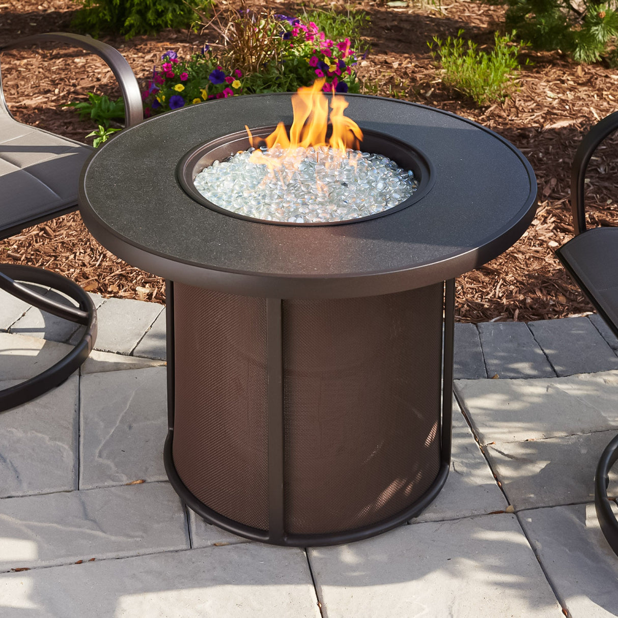 The Brown Stonefire Round Gas Fire Pit Table on a patio setup surrounded by patio furniture