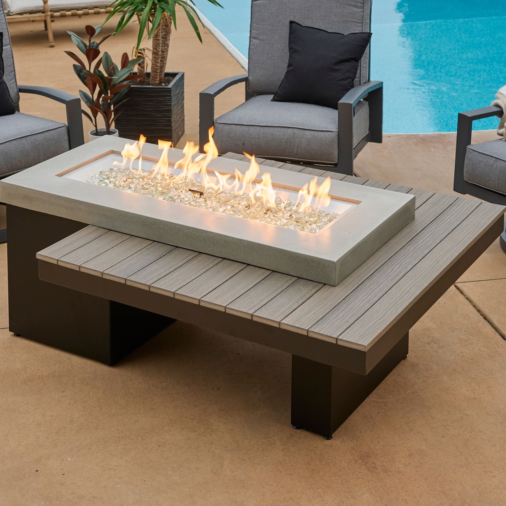 The Uptown Coastal Grey Linear Gas Fire Pit Table on a patio with a flame