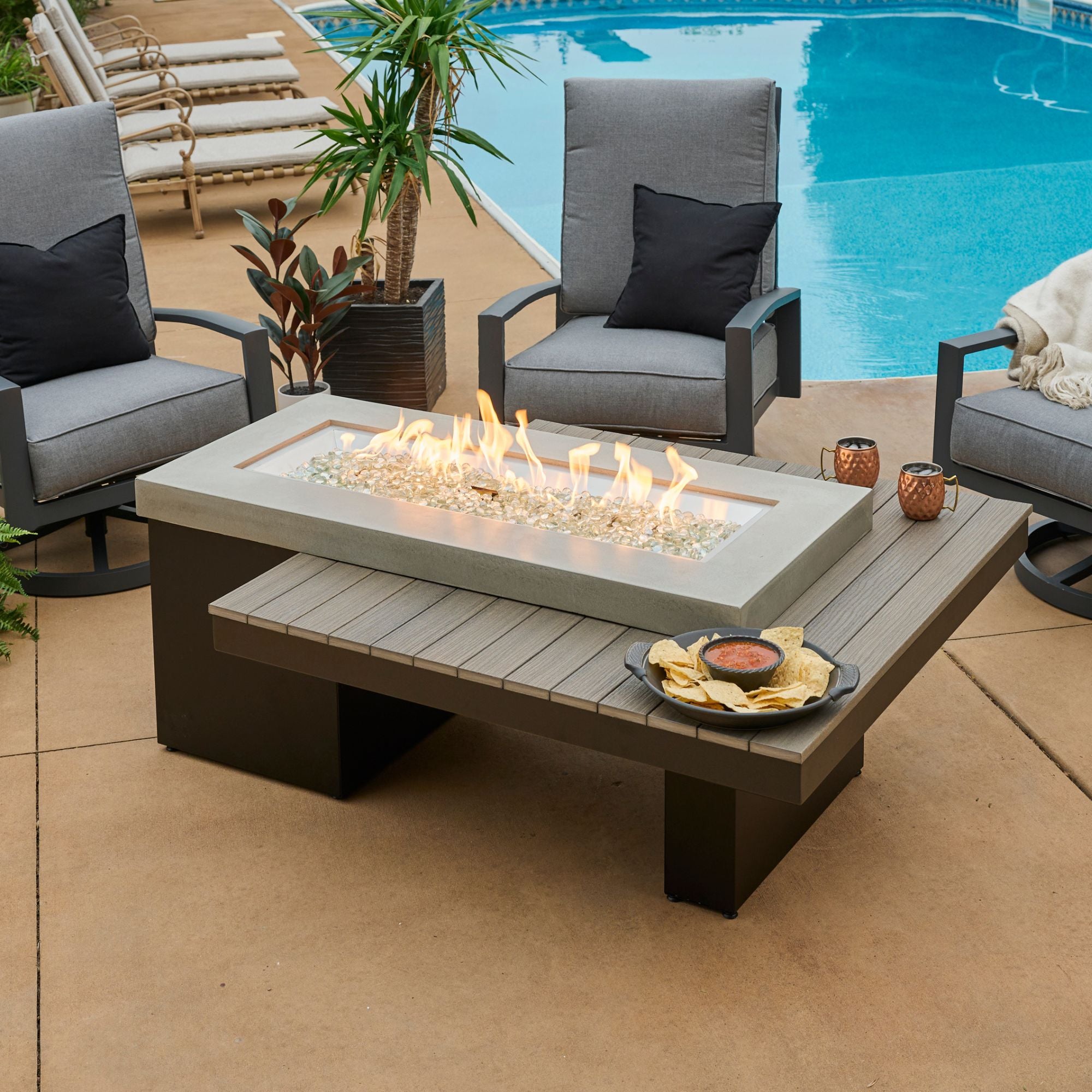 The Uptown Coastal Grey Linear Gas Fire Pit Table on a patio with a flame, drinks, and food on the tabletop 