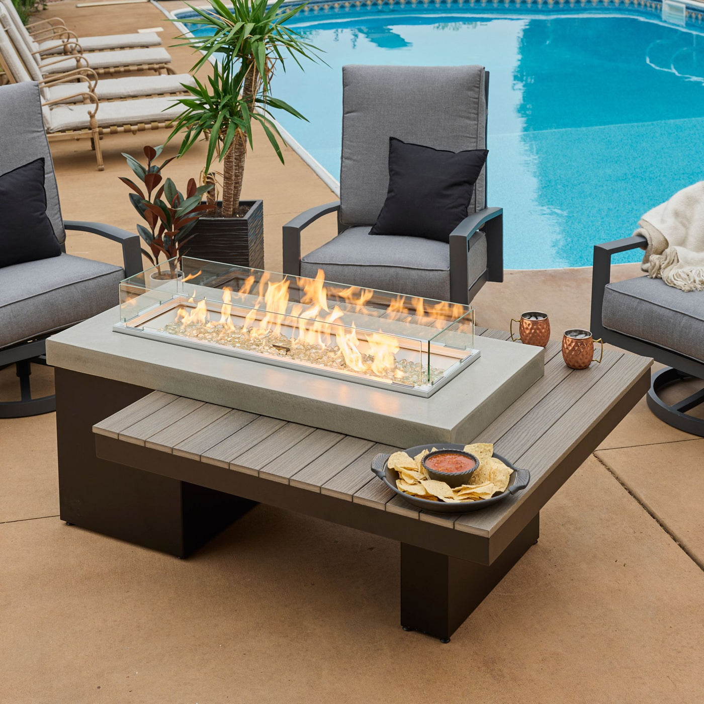 The Uptown Coastal Grey Linear Gas Fire Pit Table on a patio with a flame, glass wind guard, drinks, and food on the tabletop