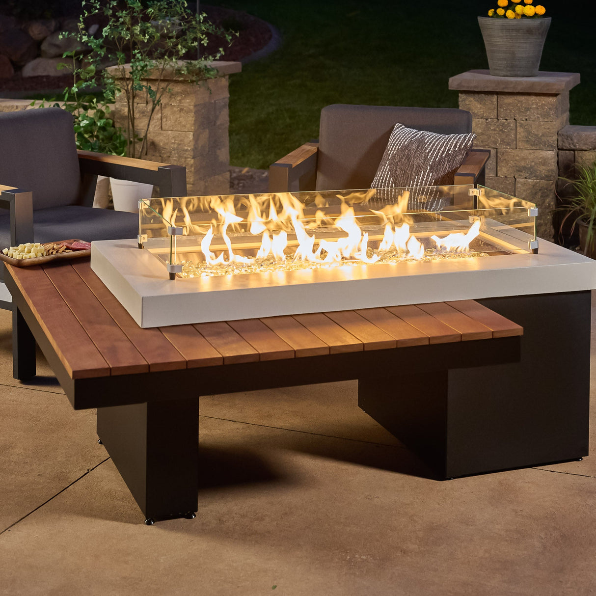 A glass wind guard placed on the top of the Uptown Iroko Linear Gas Fire Pit Table along with some food on the edge