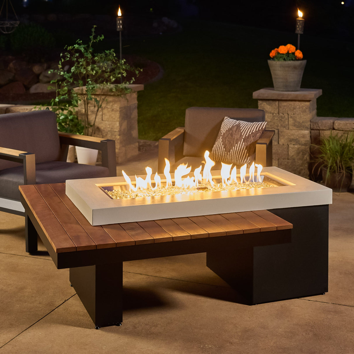 The Uptown Iroko Linear Gas Fire Pit Table on a patio with a large flame on a dark night