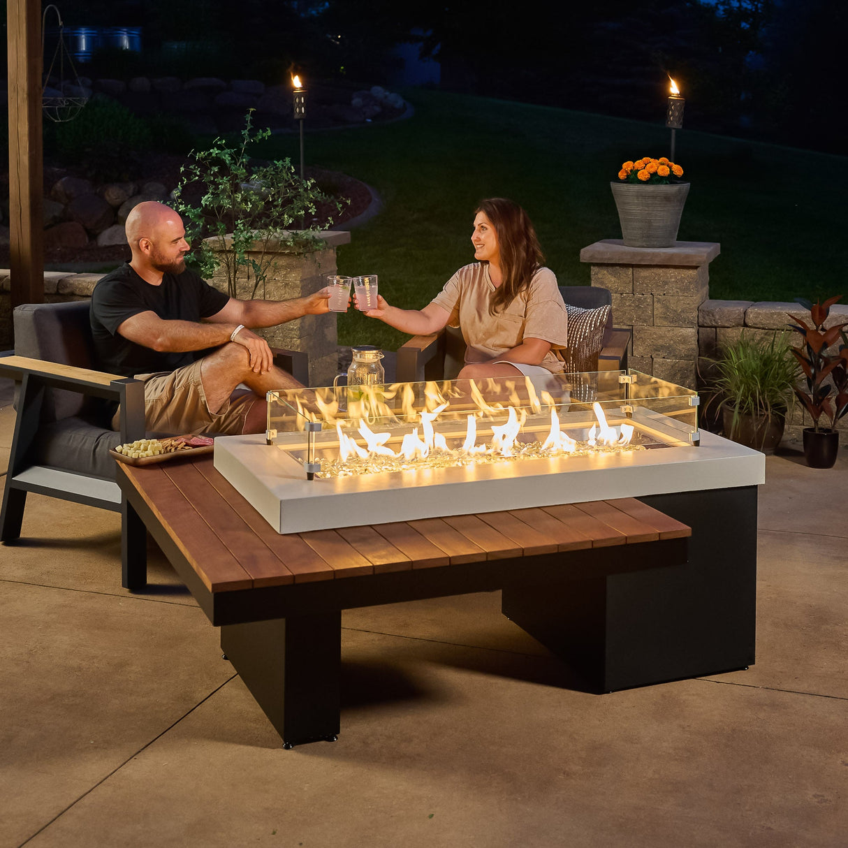 Two individuals sharing a drink and food next to the Uptown Iroko Linear Gas Fire Pit Table