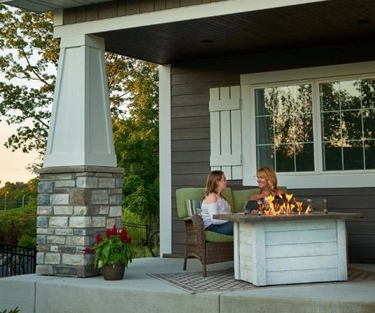 Two individuals on a back patio sitting next to an Alcott Rectangular Gas Fire Pit Table