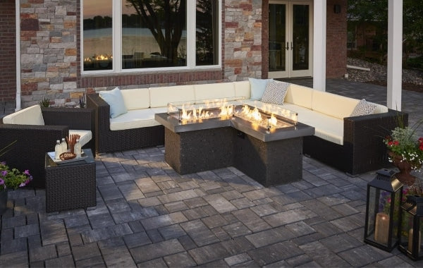 An outdoor patio setup with a large l-shaped linear gas fire pit table surrounded by wicker patio furniture